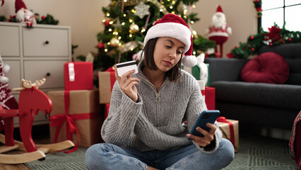 Young beautiful hispanic woman using smartphone and credit card sitting on floor by christmas tree at home
