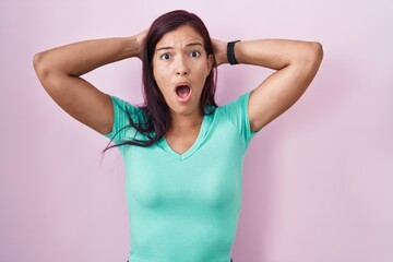 Young hispanic woman standing over pink background crazy and scared with hands on head, afraid and surprised of shock with open mouth