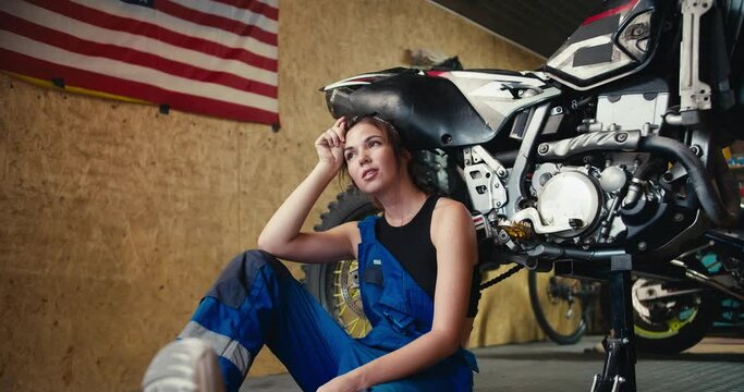 A tired girl auto mechanic in a black top and blue overalls leans on a motorcycle that she jacked up and rests in her workshop with a US flag on the wall
