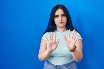 Young modern girl with blue hair standing over blue background moving away hands palms showing refusal and denial with afraid and disgusting expression. stop and forbidden.