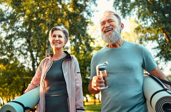 Portrait of two active sporty bright senior athletes holding exercise mats and walking outdoors in the park. Active old age. A walk, on the way to training.