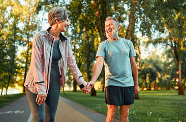 Cheerful modern senior couple outdoors in the park dressed in sportswear walking holding hands and...