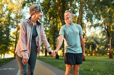 Fotobehang Cheerful modern senior couple outdoors in the park dressed in sportswear walking holding hands and laughing. Morning sun rays fall on smiling man's face.. © HBS