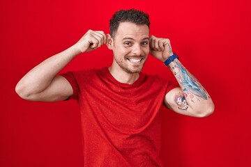 Young hispanic man standing over red background smiling pulling ears with fingers, funny gesture....