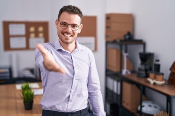 Young hispanic man at the office smiling cheerful offering palm hand giving assistance and acceptance.