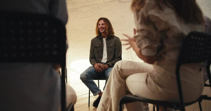 A man with curly red hair in a green gray sweater expresses his problem at groove therapy in a white brick hall. Other participants in a group therapy meeting applaud the man