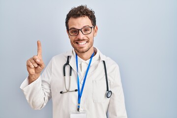 Young hispanic man wearing doctor uniform and stethoscope showing and pointing up with finger number one while smiling confident and happy.