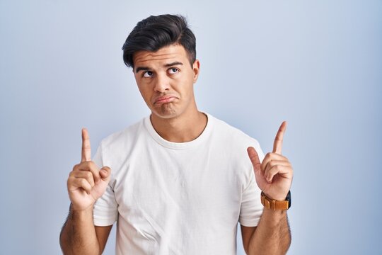 Hispanic man standing over blue background pointing up looking sad and upset, indicating direction with fingers, unhappy and depressed.