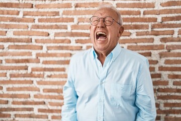 Senior man with grey hair standing over bricks wall angry and mad screaming frustrated and furious, shouting with anger. rage and aggressive concept.