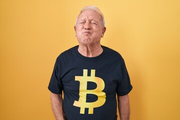 Senior man with grey hair wearing bitcoin t shirt puffing cheeks with funny face. mouth inflated...