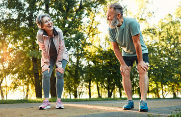 Happiness in street exercising. Senior couple feeling exhaust and taking short break to regain strength after running outside. Positive people in sport outfits laughing while bending for rest. - obrazy, fototapety, plakaty
