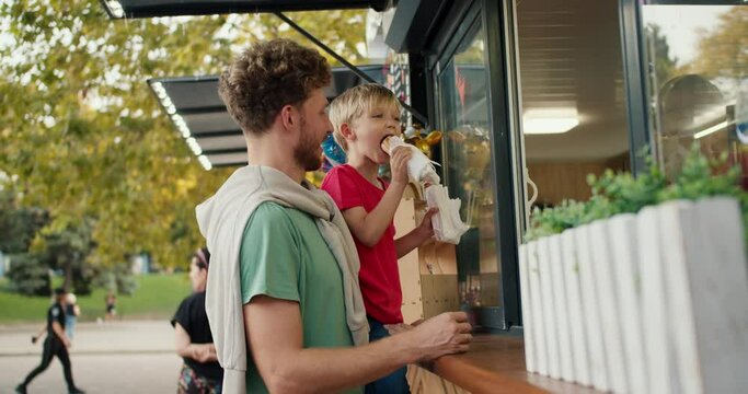 Happy little blond boy in a red T-shirt receives two hot dogs from the window of a street cafe in the park while sitting in the arms of his dad with curly hair in a Green T-shirt. Father and son