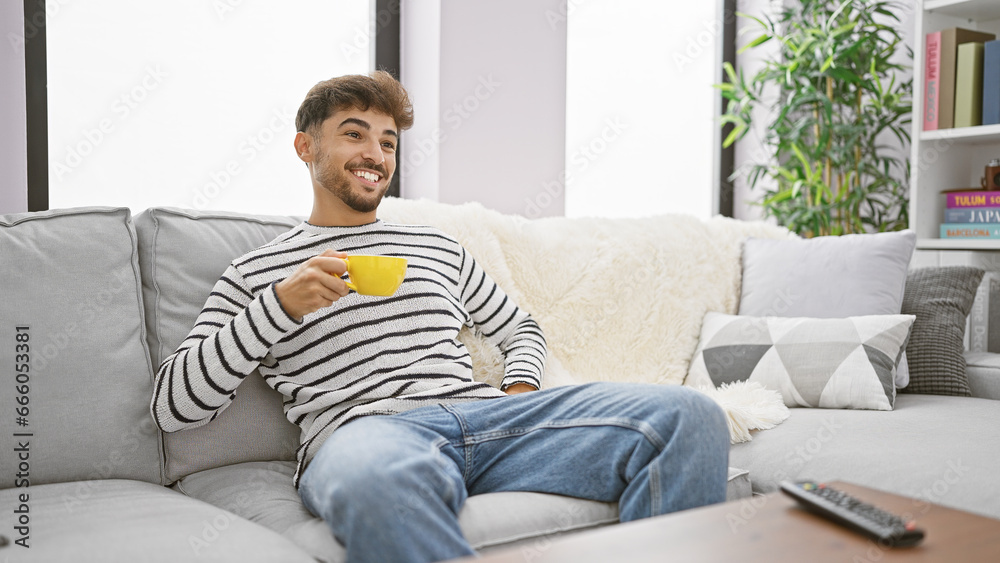 Sticker Chilled-out young arab man smiling as he enjoys his morning espresso, comfortably relaxed watching a film on his tv, living room background.