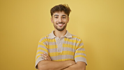 Young, smiling adult, an arab man's expression of joy and confidence, standing with folded arms isolated on a yellow background