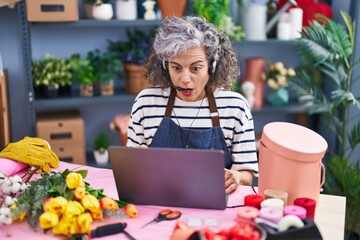 Middle age woman with grey hair working at florist with laptop scared and amazed with open mouth for surprise, disbelief face