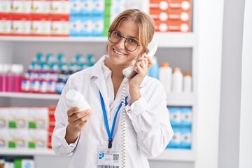 Young blonde girl pharmacist holding pills bottle talking on telephone at pharmacy
