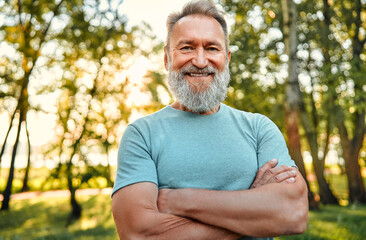 Vitality and active lifestyle. Portrait of fit mature man with hands crossed posing on camera during active training in green forest. Grey-bearded sportsman in blue shirt making confident smile. - obrazy, fototapety, plakaty