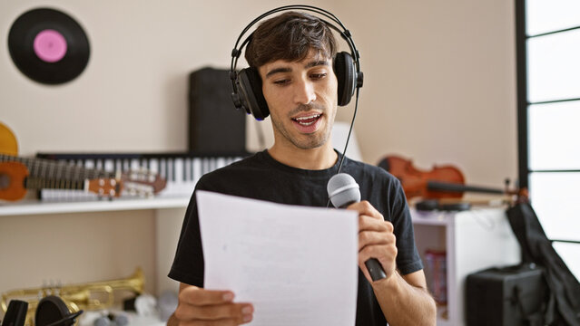Passionate young hispanic man, a handsome musician, in the heart of a melody, reading music sheet and singing the song with a professional voice at an indoor music studio.
