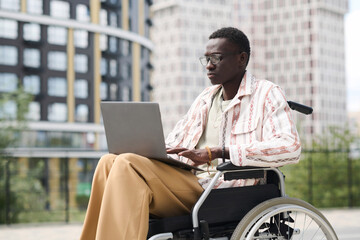 African American man with disability sitting on wheelchair and typing on laptop outdoors