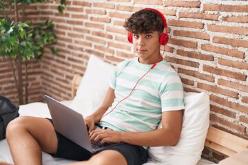 Young hispanic teenager using laptop and headphones sitting on bed at bedroom