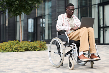 Fototapeta na wymiar African American man sitting on wheelchair and using laptop outdoors in the city