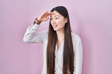 Chinese young woman standing over pink background very happy and smiling looking far away with hand over head. searching concept.