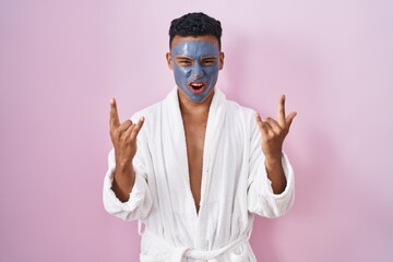 Young hispanic man wearing beauty face mask and bath robe shouting with crazy expression doing rock symbol with hands up. music star. heavy music concept.