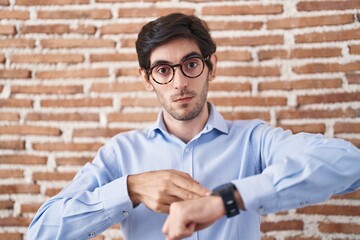 Young hispanic man standing over brick wall background in hurry pointing to watch time, impatience,...