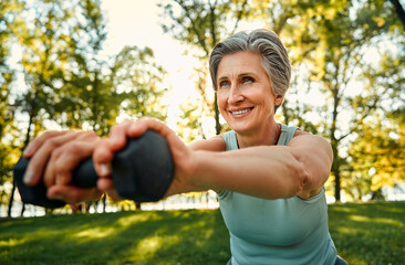 Regular workout outdoors. Close up of smiling aged lady with stylish haircut training on fresh air with dumbbells. Sporty woman strengthening muscles and bones with slight exercises. - obrazy, fototapety, plakaty