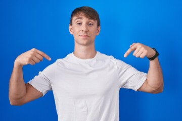 Caucasian blond man standing over blue background looking confident with smile on face, pointing...
