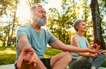 Fotobehang Physical activity on nature. Two sport partners spending morning time in park and doing yoga exercises. Active pensioners meditating in lotus pose on rugs, trying to relax mind and relieve stress. © HBS