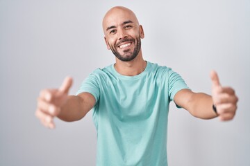 Middle age bald man standing over white background looking at the camera smiling with open arms for hug. cheerful expression embracing happiness.