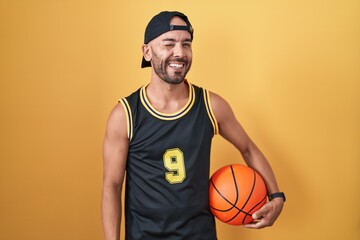 Middle age bald man holding basketball ball over yellow background winking looking at the camera with sexy expression, cheerful and happy face.
