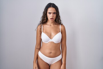 Obraz premium Young hispanic woman wearing white lingerie in shock face, looking skeptical and sarcastic, surprised with open mouth