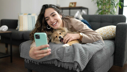 Young hispanic woman with dog taking selfie picture with smartphone lying on sofa at home - obrazy, fototapety, plakaty