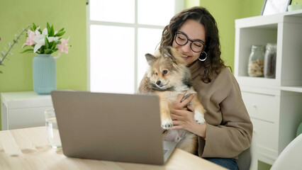 Young hispanic woman with dog sitting on table having video call at dinning room