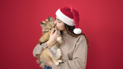 Young hispanic woman with dog wearing christmas hat kissing over isolated red background