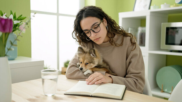 Young hispanic woman with dog sitting on table reading book at dinning room