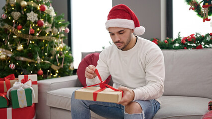 Obraz na płótnie Canvas Young hispanic man wearing christmas hat unpacking gift at home