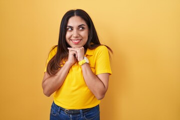 Young arab woman standing over yellow background laughing nervous and excited with hands on chin...