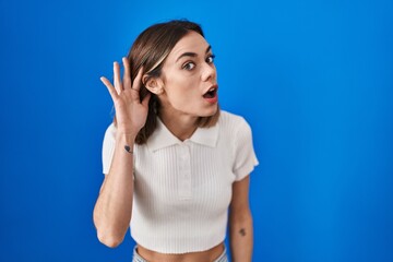 Hispanic woman standing over blue background smiling with hand over ear listening an hearing to...