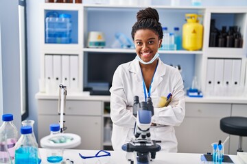 African american woman scientist wearing medical mask with arms crossed gesture at laboratory