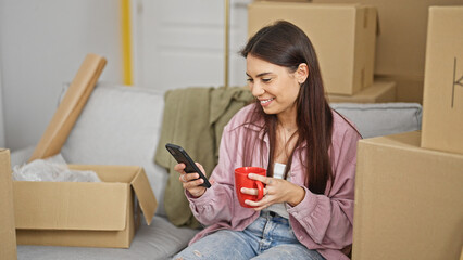 Young beautiful hispanic woman using smartphone drinking coffee at new home
