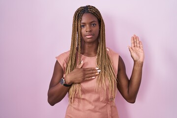 African american woman with braided hair standing over pink background swearing with hand on chest...
