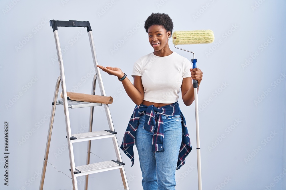 Wall mural African american woman holding roller painter pointing aside with hands open palms showing copy space, presenting advertisement smiling excited happy