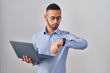 Young hispanic man working using computer laptop checking the time on wrist watch, relaxed and confident