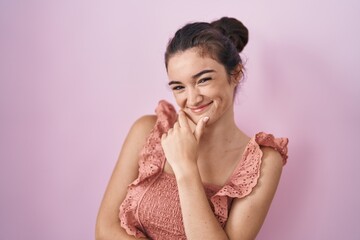 Young teenager girl standing over pink background looking confident at the camera smiling with crossed arms and hand raised on chin. thinking positive.
