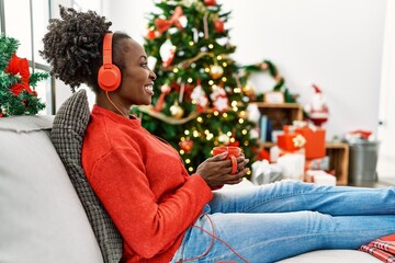 Obraz na płótnie Canvas African american woman listening to music drinking coffee sitting on sofa by christmas tree at home