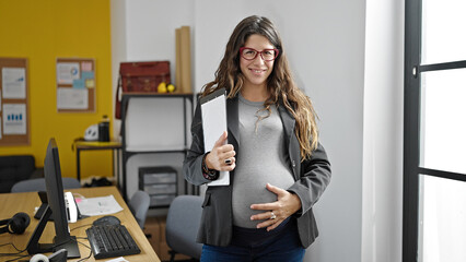 Young pregnant woman business worker touching belly holding documents at office