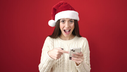 Young beautiful hispanic woman wearing christmas hat pointing with finger to smartphone over isolated red background
